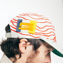 Load image into Gallery viewer, A close-up side profile of a man wearing a white baseball cap with red wavy stripes, a blue and yellow embroidered patch, and a green brim, paired with a beige jacket against a plain white background.
