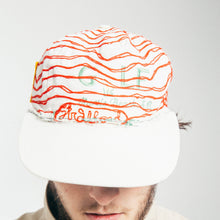 Load image into Gallery viewer, A man wearing a white baseball cap with orange wavy stripes and colorful embroidered text, paired with a beige high-collar jacket, against a light gray background.

