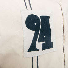 Load image into Gallery viewer, Close-up of a beige jacket with a large black '94' patch on the chest, dark double-stripe detailing, and a buttoned collar.
